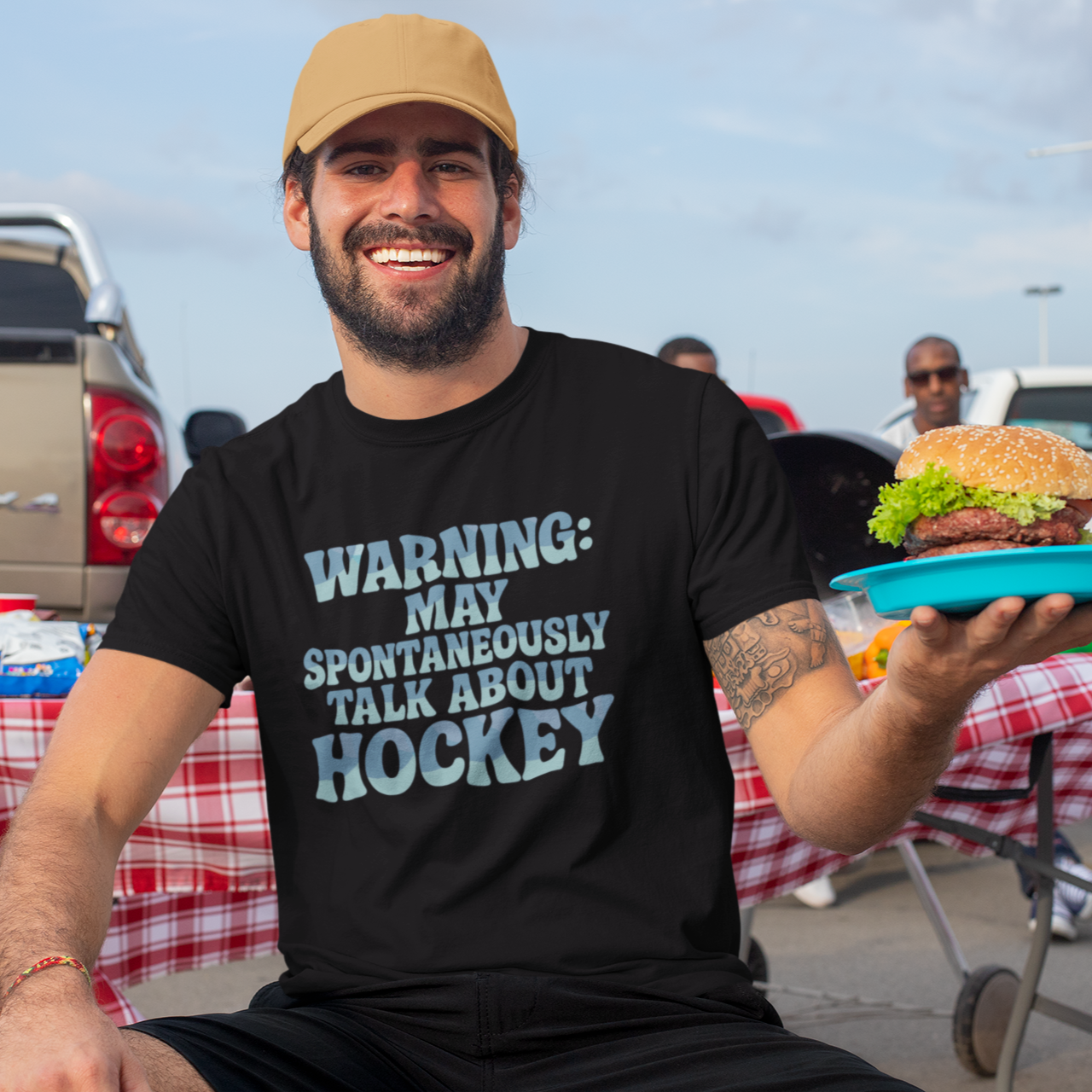 Man holding a burger in a parking lot wearing a black t-shirt colorful text "Warning: may spontaneously talk about hockey"